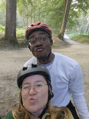 Dos amigos con cascos y gafas posan para un juguetón selfie en un bosque, haciéndose carantoñas. El fondo muestra altos árboles y un camino de tierra.