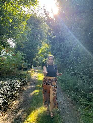 Una mujer de pie en un camino forestal iluminado por el sol, rodeada de exuberante vegetación. Los rayos del sol se cuelan entre los árboles, creando un ambiente tranquilo y relajado. Gesticula abiertamente.