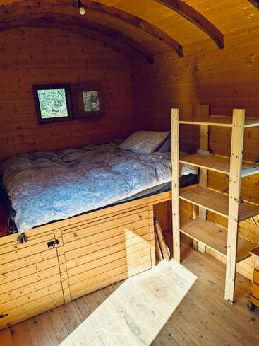 Das Bild zeigt eine gemütliche Holzhütte mit einem Bett, einer Leiter und einem Fenster, die ein ruhiges und rustikales Interieur mit natürlichem Lichteinfall bietet.