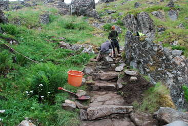 Zwei Personen bauen oder reparieren einen Steinweg in einer felsigen, grasbewachsenen Landschaft. Werkzeuge und ein orangefarbener Eimer stehen in der Nähe. Wildblumen und Farne umgeben die Szene.