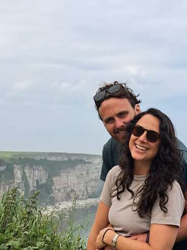 A imagem mostra um homem e uma mulher posando para uma foto em frente a um penhasco, com os longos cabelos escuros e a camiseta cinza da mulher em destaque, e o homem atrás dela.