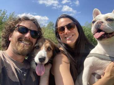 A imagem mostra um homem e uma mulher posando com dois cães, em um cenário de árvores e um céu azul com nuvens brancas.