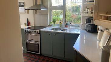 Una cocina compacta y moderna con armarios azules, horno de acero inoxidable, fregadero bajo la ventana, diversos electrodomésticos y suelo a cuadros. La luz del sol ilumina el espacio, resaltando la vegetación del exterior.