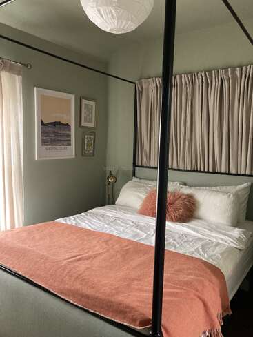 Un acogedor dormitorio cuenta con una cama con dosel con sábanas blancas, una manta rosa, una almohada mullida, cortinas suaves, cuadros en las paredes de color verde salvia y una suave luz natural.