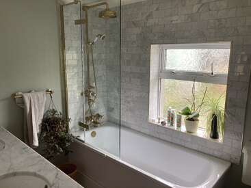Un cuarto de baño moderno con encimera de mármol, grifería dorada, mampara de ducha de cristal y bañera blanca. En el alféizar de una ventana esmerilada hay plantas y artículos de aseo.