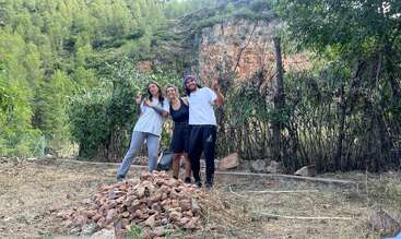 Tres personas posan sonrientes al aire libre con signos de la paz junto a un montón de rocas, rodeadas de árboles y montañas, disfrutando de la naturaleza y la camaradería en un entorno rústico.
