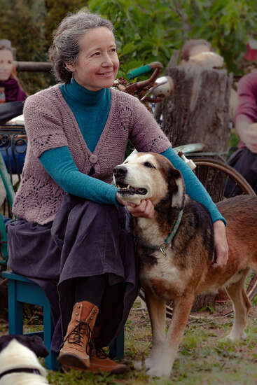 Une femme souriante aux cheveux gris est assise à l'extérieur, caressant affectueusement son gros chien. Elle porte un pull tricoté, une jupe longue et des bottes. De la verdure les entoure.