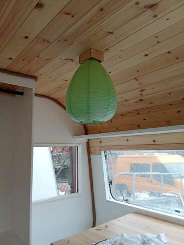 Cette pièce confortable au plafond en bois est ornée d'une lanterne en papier verte en forme de goutte d'eau. Les fenêtres lumineuses révèlent une camionnette orange à l'extérieur, tandis que la lumière naturelle remplit l'espace intérieur simple et accueillant.