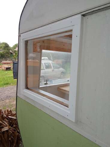 L'image montre une fenêtre rectangulaire à cadre blanc installée sur le côté d'un camping-car ou d'une caravane vert clair et blanc, avec une camionnette à l'extérieur.