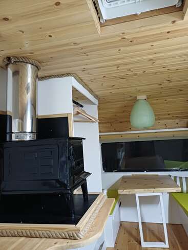 Das gemütliche Interieur des Wohnmobils besticht durch Holzvertäfelungen, einen kleinen schwarzen Holzofen, eine grüne Papierlaterne, eine kompakte Essecke mit Bänken und einen Bereich zum Aufhängen von Kleidung.
