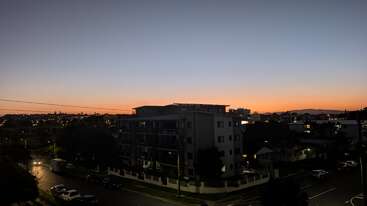 La imagen capta un barrio urbano al atardecer. Las luces de la ciudad brillan, se ven edificios y árboles en silueta, y un horizonte de color naranja marca la puesta de sol que se desvanece.