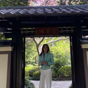 Una mujer sonríe bajo una puerta de madera tradicional japonesa, rodeada de frondosos jardines y árboles. Lleva una chaqueta verde azulado y pantalones blancos, y sostiene su teléfono.