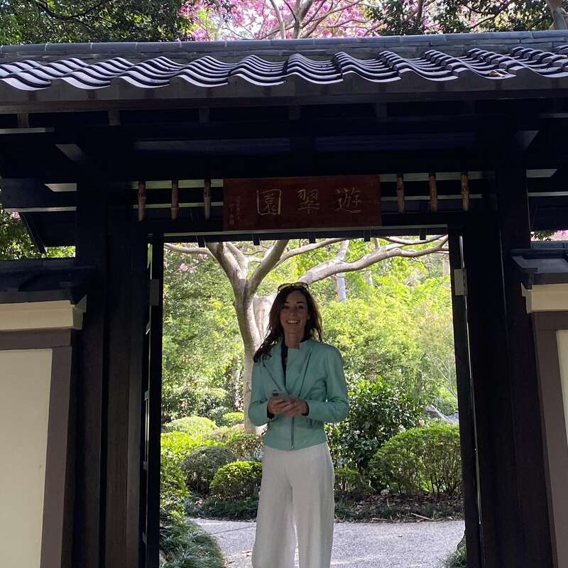 Una mujer sonríe bajo una puerta de madera tradicional japonesa, rodeada de frondosos jardines y árboles. Lleva una chaqueta verde azulado y pantalones blancos, y sostiene su teléfono.