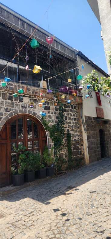 Uma rua de paralelepípedos ao lado de um prédio de pedra com portas de madeira em arco, vasos de plantas e casinhas de pássaros coloridas penduradas acima, sob um céu azul claro.