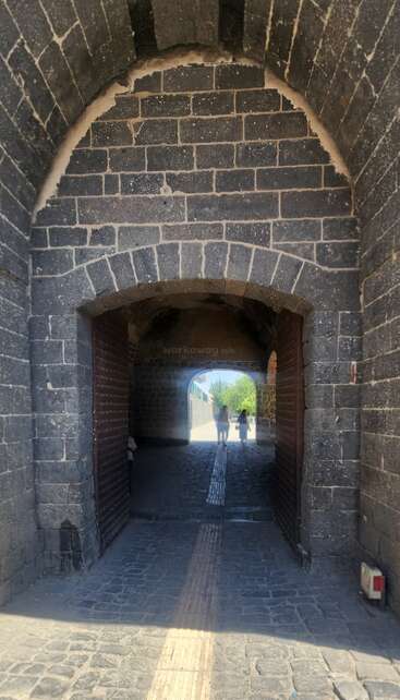 Esta imagem mostra um túnel em arco de pedra com duas pessoas caminhando em direção à saída. A luz do sol ilumina o caminho de paralelepípedos e a vegetação do lado de fora do túnel.