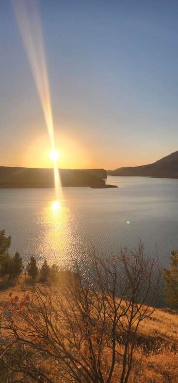 A luz dourada do sol reflete em um lago calmo, com grama seca e arbustos em primeiro plano. Colinas distantes e céu claro completam essa paisagem tranquila do pôr do sol.