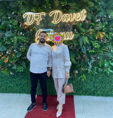 Um casal está de mãos dadas em um tapete vermelho diante de um cenário de folhas verdes e do letreiro em neon "DF Davet Salonu". O rosto da mulher está coberto.