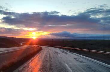 Uma estrada sinuosa se estende ao longe, refletindo os tons vibrantes de laranja e rosa de um pôr do sol de tirar o fôlego, com nuvens dramáticas preenchendo o céu acima.