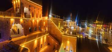 Esta imagem mostra um edifício histórico maravilhosamente iluminado à noite, adornado com luzes amarelas quentes. A arquitetura apresenta arcos, escadas e vários detalhes intrincados.