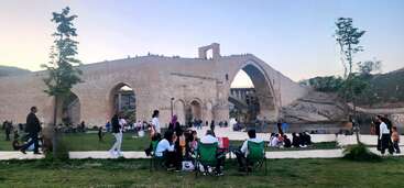 Uma ponte histórica de pedra atravessa um rio, com muitas pessoas reunidas embaixo e ao redor dela, desfrutando de um piquenique em um parque gramado durante um dia agradável.