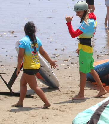 Dos personas con camisetas de colores y ropa de playa están de pie en la orilla arenosa, una lleva una tabla de surf con un hidrofoil, mientras que la otra hace el signo shaka.
