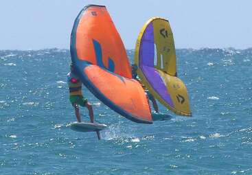 Dos personas practican wing foil en el océano. Una tiene un ala naranja y la otra, una ala amarilla y morada. Ambas se deslizan sobre el agua.