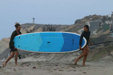 Dos personas llevan alegremente una gran tabla de paddle surf por una playa de arena, con acantilados rocosos y un faro visibles al fondo, bajo un cielo nublado.
