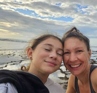 A woman and a girl are smiling together for a selfie at the beach. The sky is cloudy, and the ocean water creates a serene backdrop.