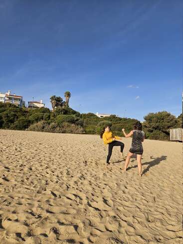Two women are playfully sparring on a sandy beach under a clear blue sky, with green bushes and houses visible in the background, enjoying a sunny day.
