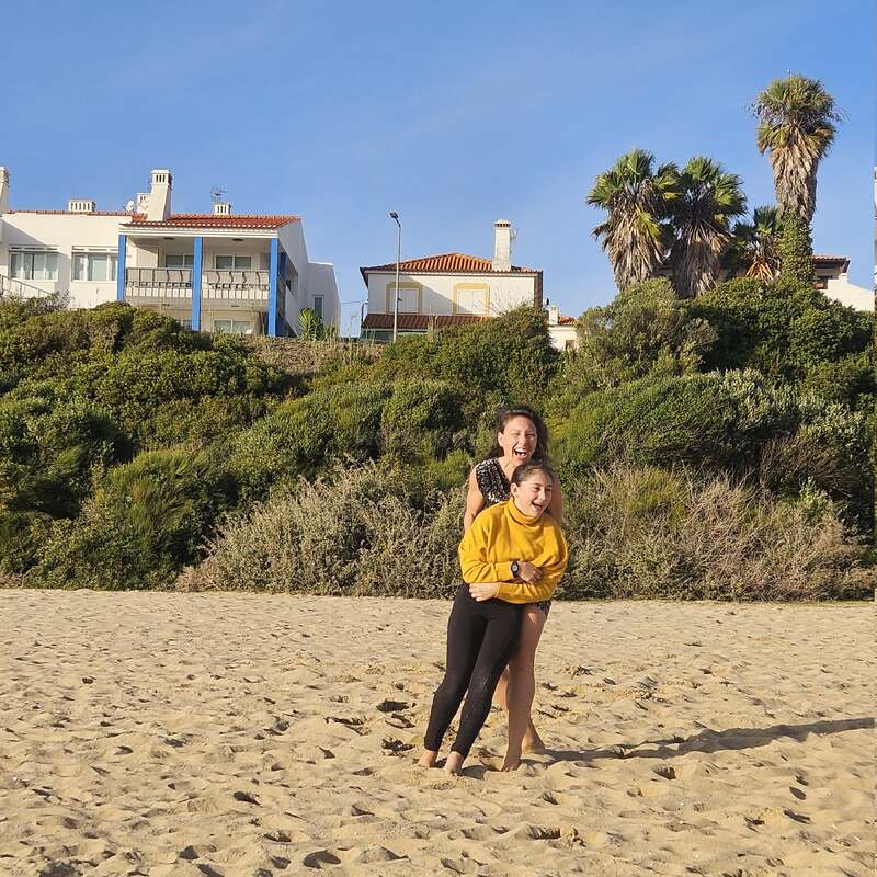 Two girls are playfully hugging and laughing on a sandy beach. Behind them are green bushes, palm trees, and white houses under a clear blue sky.