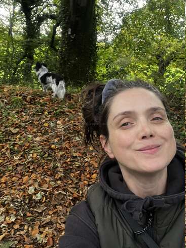 Eine Frau macht ein Selfie in einem Waldgebiet und lächelt dabei herzlich. Hinter ihr steht ein schwarz-weißer Hund auf einem mit Blättern bedeckten Boden, umgeben von Bäumen.