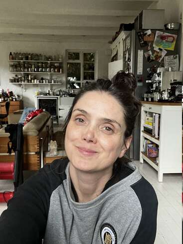 Eine Frau mit entspanntem Gesichtsausdruck macht ein Selfie in einer gemütlichen, wohnlichen Küche. Regale voller Gläser, ein Kühlschrank mit Magneten und warmes Tageslicht.