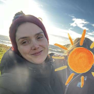 Uma mulher com um gorro vermelho sorri ao ar livre, perto do mar, vestindo uma jaqueta escura. O sol brilha forte e um desenho de sol cobre o rosto de outra pessoa.