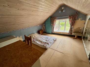 Esta imagen muestra una habitación en el ático con escaso mobiliario, techo inclinado de madera, cama individual, escritorio, silla, una ventana grande con cortinas de flores y paredes y suelo parcialmente sin terminar.