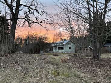 Una casa rústica se alza entre árboles sin hojas al atardecer. El cielo resplandece en tonos anaranjados y rosados, mientras un perro peludo pasea por el camino del jardín.