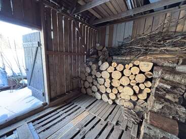 La luz del sol inunda un cobertizo rústico de madera con la puerta abierta, leña apilada con cuidado, ramas atadas en fardos, una cesta metálica y, al exterior, un paisaje cubierto de nieve. Ambiente acogedor y rural.