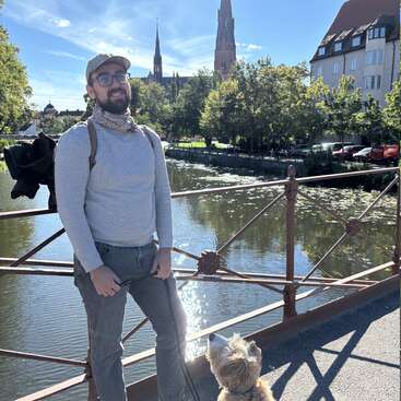 Um homem sorridente, de óculos e boné, está em uma ponte segurando a coleira de um cachorro. Fundo ensolarado, com rio, árvores, catedral e prédios da cidade.