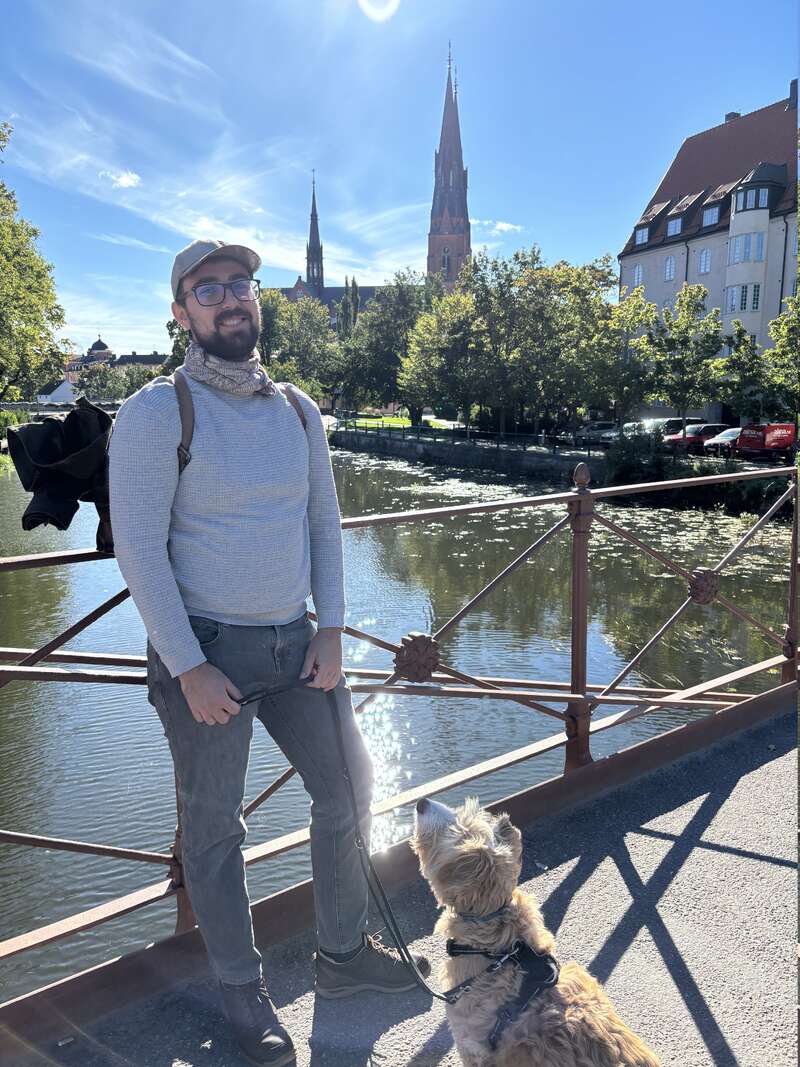 Un hombre sonriente, con gafas y gorra, está de pie en un puente sujetando la correa de un perro. Al fondo se ven un día soleado, un río, árboles, una catedral y los edificios de la ciudad.