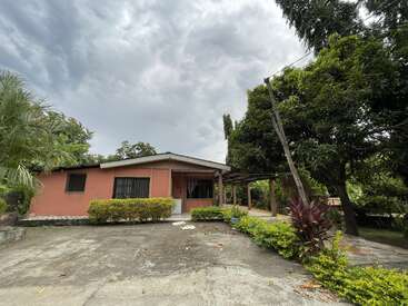 La imagen muestra una pequeña casa de una planta con cochera, rodeada de frondosos árboles y un cielo nublado.