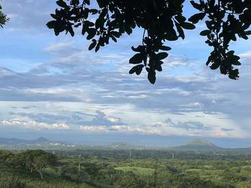 La imagen representa un paisaje sereno con un frondoso valle verde, montañas lejanas y un cielo azul con nubes blancas, enmarcado por ramas de árboles en la parte superior.