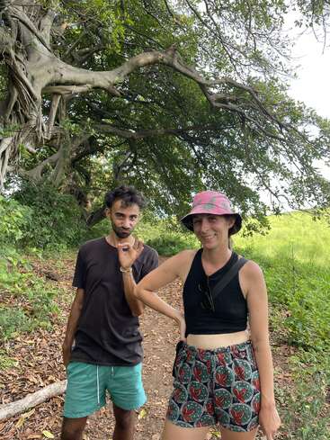 La imagen muestra a un hombre y una mujer de pie en un camino de tierra frente a un árbol, con la mujer haciendo un gesto de "okay" con la mano.