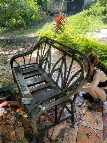 Una mujer está agachada en un patio, fregando con un cepillo un banco de madera desgastada, rodeada de plantas y árboles en un entorno sereno al aire libre.