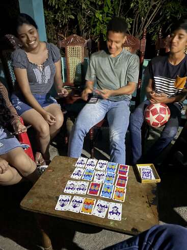 La imagen muestra a un grupo de cuatro personas reunidas en torno a una mesa de madera, jugando a las cartas en un ambiente nocturno al aire libre. El ambiente parece relajado y agradable.