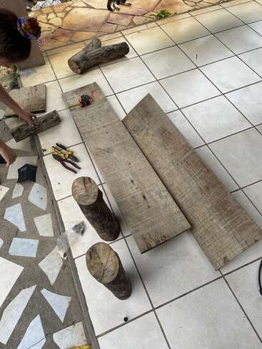 La imagen muestra a una mujer colocando tablones y troncos de madera sobre un suelo de baldosas, rodeada de herramientas y escombros, con un perro visible al fondo.