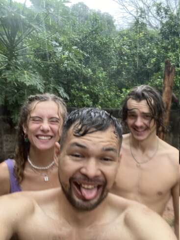 La imagen muestra a tres personas posando para un selfie bajo la lluvia, con exuberante vegetación y árboles de fondo, rodeados por una valla de madera.