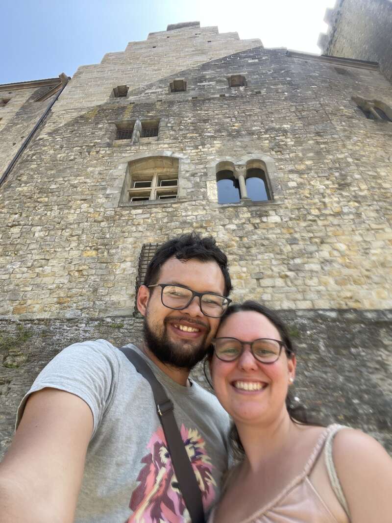 La imagen muestra un selfie de un hombre y una mujer frente a un antiguo edificio de piedra con una torre y ventanas, enmarcado en un cielo azul.