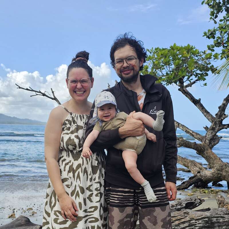 Eine lächelnde dreiköpfige Familie - Mutter, Vater und Baby - posiert glücklich an einem tropischen Strand, mit Bäumen, blauem Himmel und Meereswellen im Hintergrund.