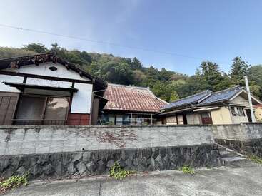 Três casas tradicionais japonesas ficam atrás de um muro de pedra. Colinas com florestas verdes se erguem ao fundo sob um céu azul claro, criando uma atmosfera pacífica e rural.