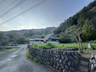 Uma casa rural com um telhado tradicional fica entre árvores verdes e colinas. Muros de pedra margeiam a estrada e linhas de energia elétrica se estendem pelo céu acima de você.
