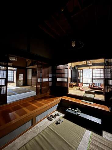 Esta imagem mostra uma casa tradicional japonesa com tapetes de tatame, interiores de madeira, portas shoji deslizantes, uma entrada genkan e chinelos arrumados na entrada. Atmosfera tranquila.